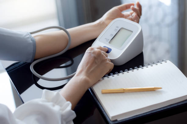Woman measuring blood pressure by using digital sphygmomanometer with empty white notebook or diary on the desk at home. Medical and healthcare concept.