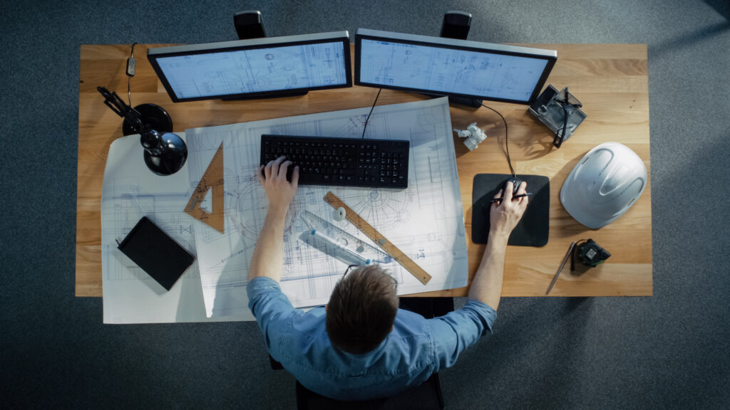 Top View of a Technical Engineer Working on His Blueprints, Drawing Plans, Using Desktop Computer. Various Useful Items Lying on his Table.