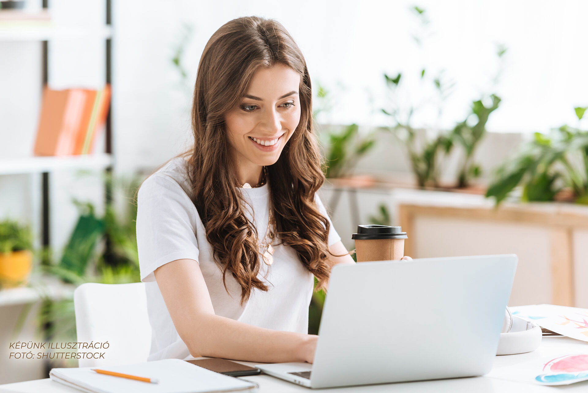 cheerful young woman using laptop and holding paper cup while sitting at desk at home