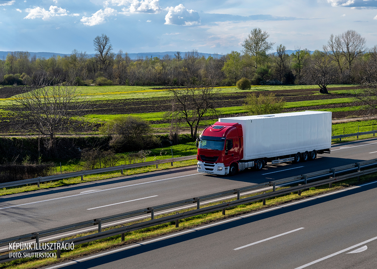 A big red truck with a white trailer drives on the highway through the rural areas of Serbia. Beautiful blue sky with white clouds above the road.